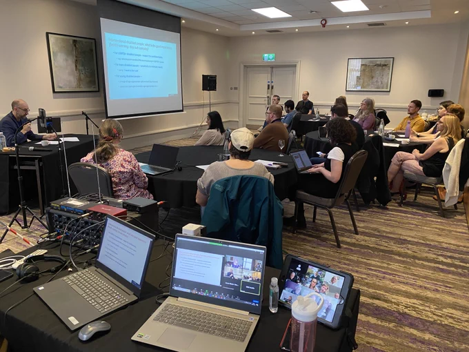 GDA members fill a room, with the hybrid set-up visible in the foreground, showing GDA members participating on Zoom.