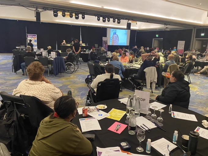 GDA members fill a ballroom. On a screen at the front of the room, a GDA member on Zoom is being projected, speaking to the room.