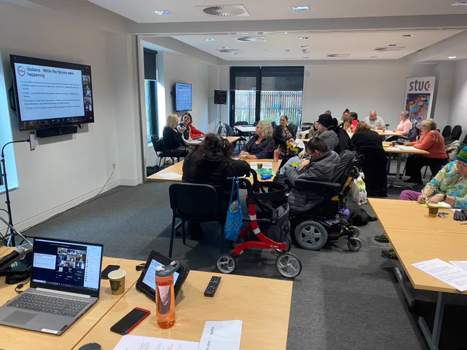 GDA members fill a room. On the wall, TV screens show PowerPoint slides and GDA members participating on Zoom. In the foreground, a laptop shows the Zoom meeting on a laptop and iPad.