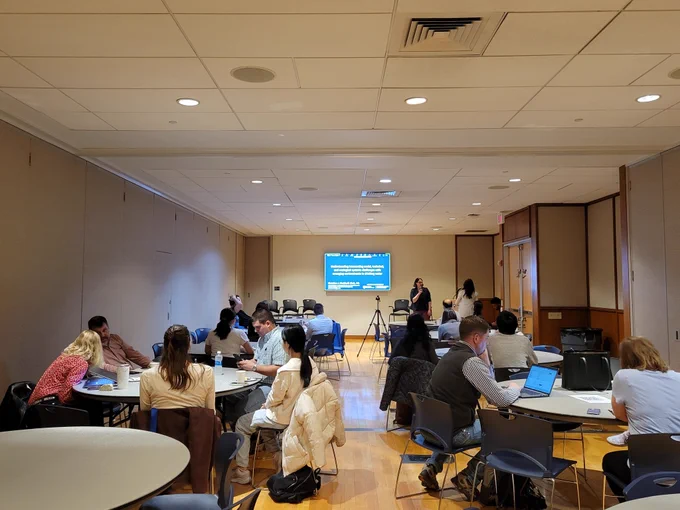 Dr. Christine Kirchoff presenting her presentation on emerging contaminants. The image shows Christine by a PowerPoint slide at the front of a room with tables surrounded by people. There are about 20 people in the room. 
