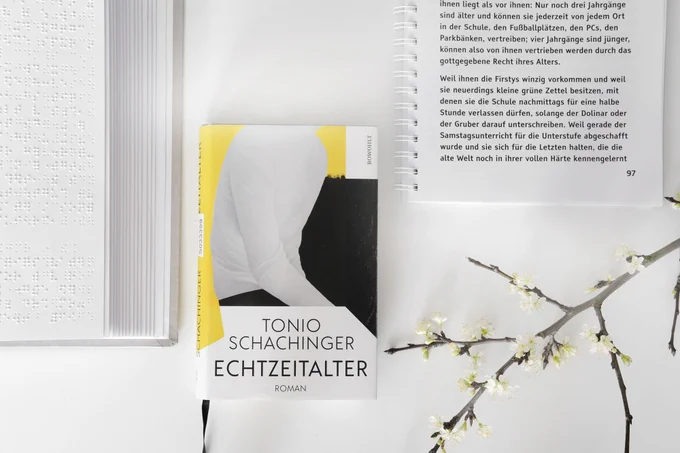 Originalbuch "Echtzeitalter" von Tonio Schachinger, links aufgeschlagenes Braillebuch (angeschnitten), rechts oben aufgeschlagenes Großdruckbuch (angeschnitten), rechts unten Blütenzweig