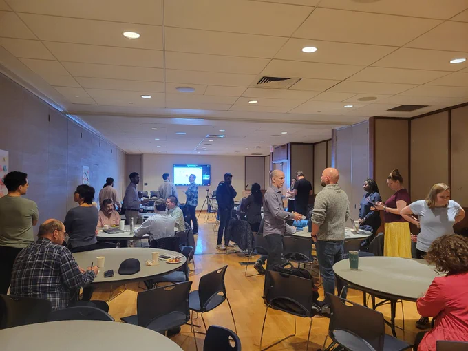 Photo of the room where people are having discussions around tables and adding sticky notes to the posters on the walls for various topics. 
