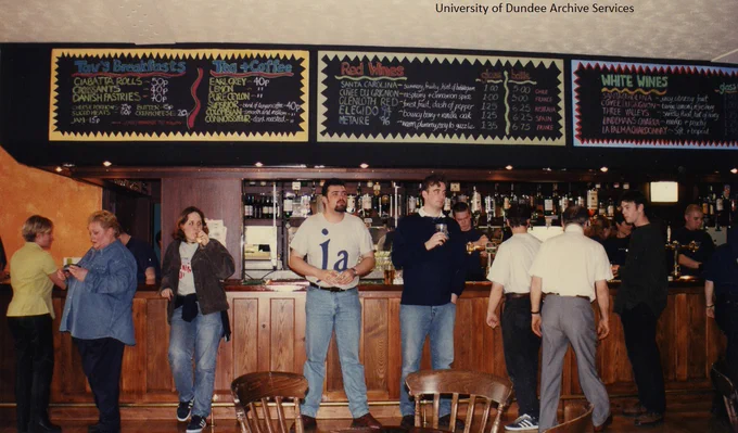 Colour photograph. A group of people standing in front of a bar, some holding drinks. Prices and products are written in coloured chalk on blackboards above the bar.