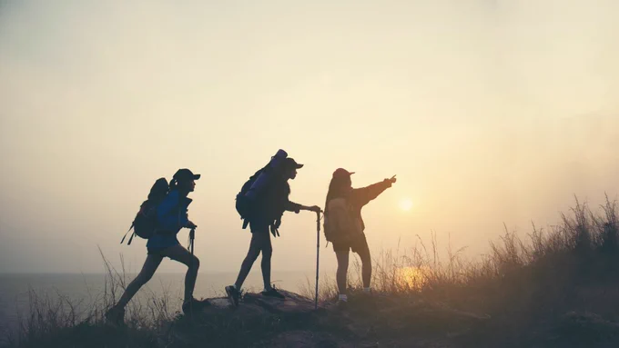 Image of 3 people hiking along a hill, the person in the front is pointing toward the right side of the image. 