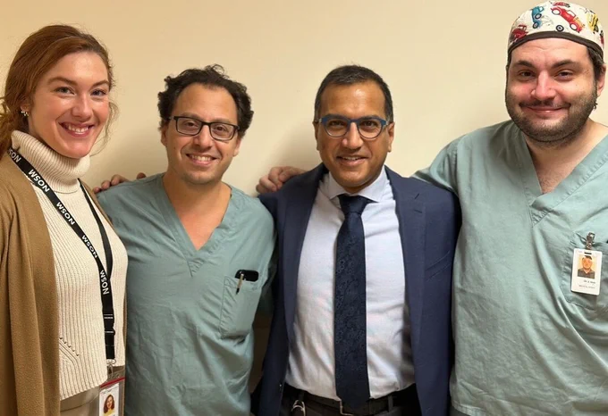 Four health-care professionals pose for a photo. Two male physicians wear scrubs, while a third wears a navy suit. A female medical student, left, wears a sweater, cardigan, and ID badge. 

Quatre professionnels de la santé posent pour une photo. Deux médecins masculins portent une blouse, tandis que le troisième est vêtu d’un costume bleu marine. Une étudiante en médecine, à gauche, porte un pull, un gilet et un badge d’identification. 