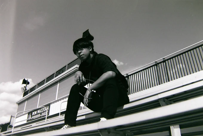 Black and white photo of Sean sitting on bleachers, press box and sky behind him. He is looking past the camera, eyes partially hidden under his bangs.