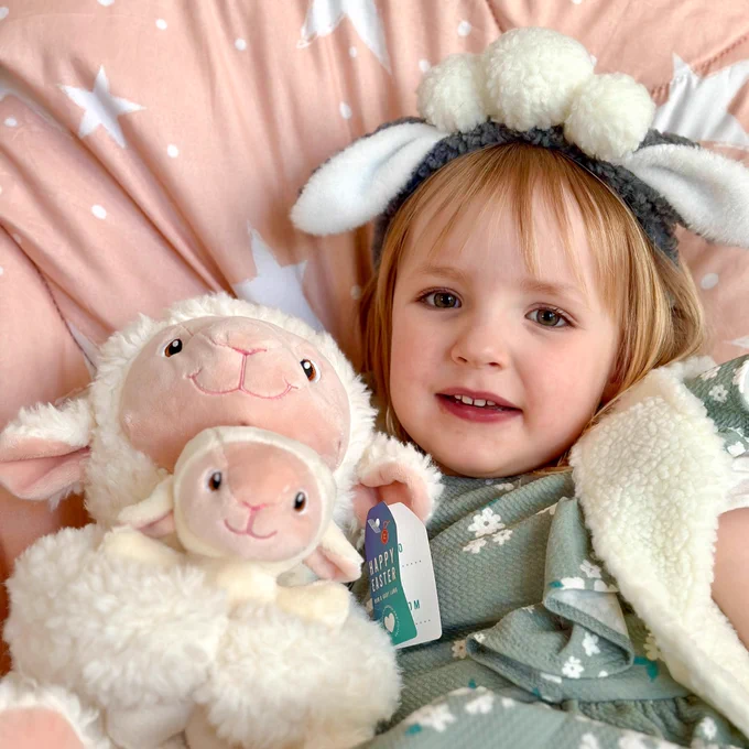 Photo shows 3.5 year old Mimi on a bed, cuddling a soft toy of a sheep with a lamb. Mimi is wearing a green dress, fluffy cream gilet and sheep ears headband