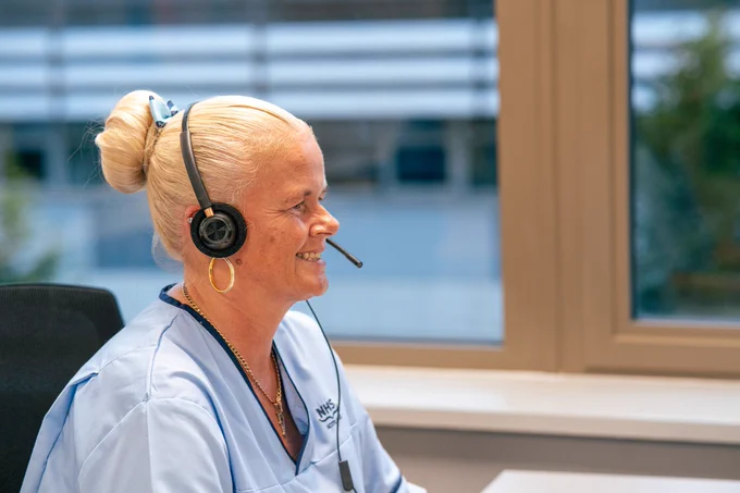 Photo of a female NHS 24 call handler. She is sitting at a desk and wearing a telephone headset. 