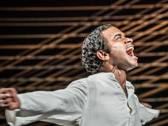 A man in a white tunic shouts in victory with his arms wide. A wall of thread behind him.