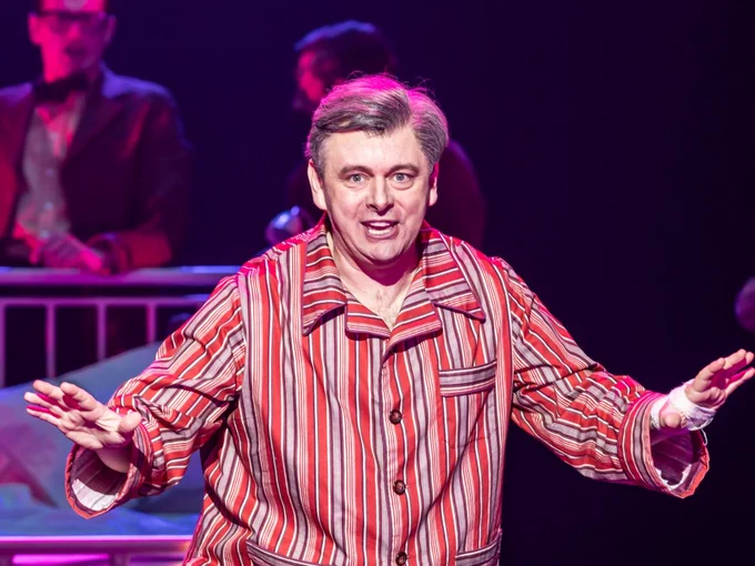 A man in red striped pyjamas sings with his arms wide. A hospital bed behind him.