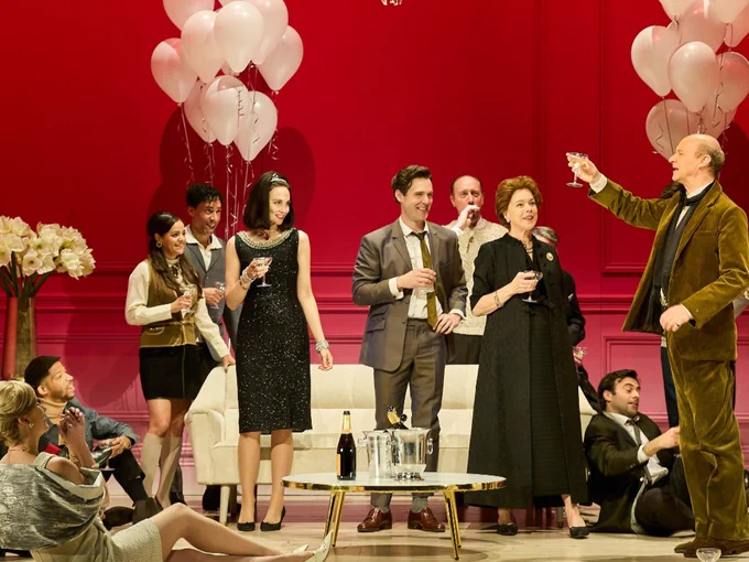 A glamourous cocktail party in the 1960s in a red painted room with balloons and a gold chandelier. A man in a green suit makes a toast and raises a coupe of champagne.