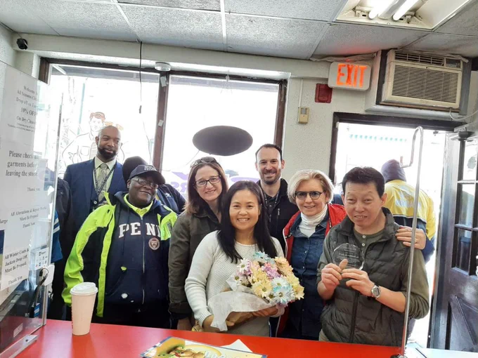 Several people pose with the owners of a local clothing cleaner business, the owners are holding flowers, and a plaque thanking them for being a part of our community.