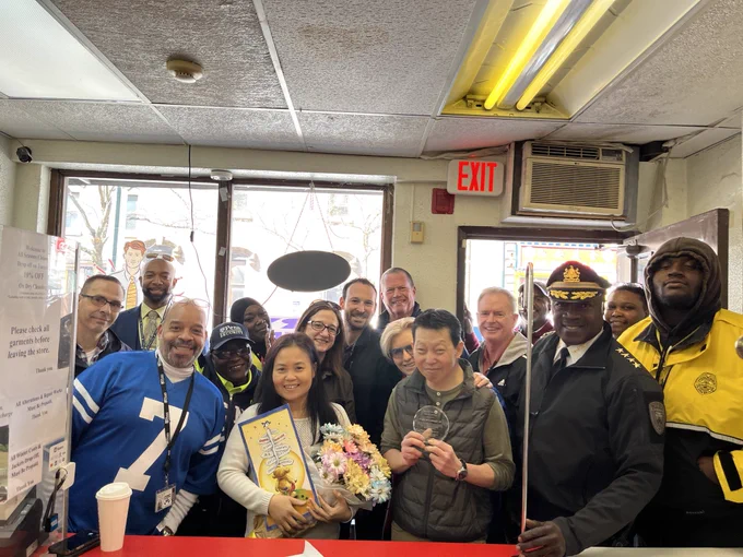 A Group of people pose with the owners in a clothing cleaner shop, the owners are holding flowers, a card, and a plaque thanking them for their partnership and friendship in the community.