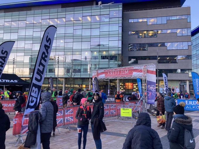 Race start area with people waiting around, flags that say GB Ultras on them and Erdinger Alkoholfrei. Modern buildings in background