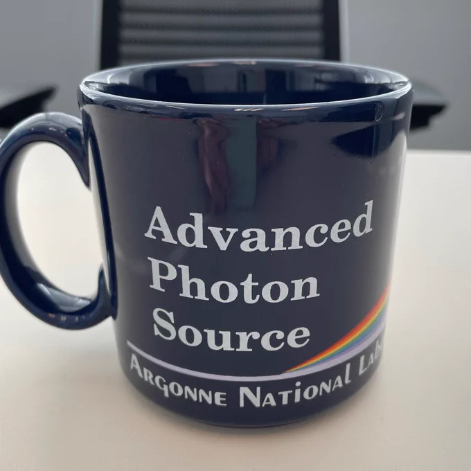 A dark blue mug with reads "Advanced Photon Source Argonne National Lab" in white text. A white line splits into a rainbow like light refracted from a prism (or a pink flloyd album cover). 