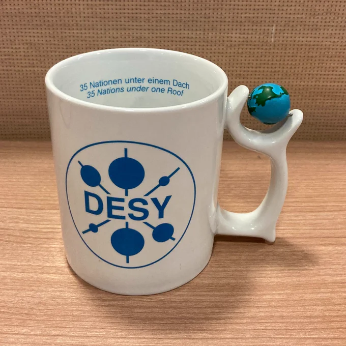 A white mug with the DESY (Deutsches Elektronen-Synchrotron) logo. Blue text inside the mug reads 35 Nationen unter einem dach and 35 nations under one roof. Affixed to the handle is a blue and green globe which can spin. 