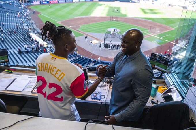 Rashid Shaheed standing in one of the Padres' broadcast booths with Tony Gwynn Jr. Rashid and Tony are smiling and dapping each other up. Rashid is wearing a custom Padres City Connect jersey that has his last name on the back (Shaheed) and the number 22 (the same number he wears on the Saints); Tony is wearing a blue button up shirt.