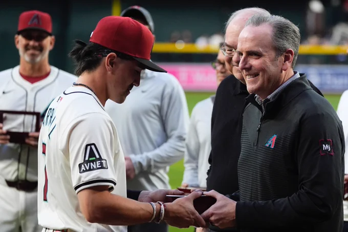 Corbin Carroll (left) receives his National League Championship ring from Arizona Diamondbacks team president Derrick Hall (right). Carroll wears a home white Arizona Diamondbacks uniform and brick red Arizona Diamondbacks hat. Derrick Hall wears an Arizona Diamondbacks quarter-zip and is smiling.