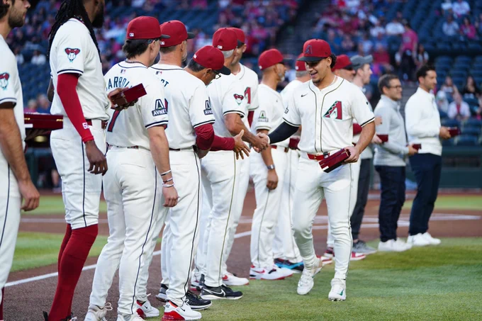 Alek Thomas greets his teammates on the baseline while holding a box containing his 2023 National League Championship ring. Thomas and the Diamondbacks all wear home white Arizona Diamondbacks uniforms and brick red Arizona Diamondbacks hats