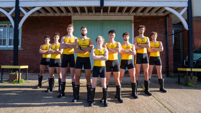 Men's reserve crew Goldie, in their bright yellow tops and black bottoms, standing in v-formation, arms crossed, in front of Goldie boathouse