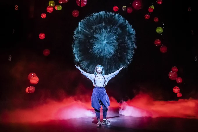 Rosie Sheehy (Puck) standing side-on and centre stage, looking up at red lit-up lanterns. Puck has her arms wide open with palms upwards at head height. Smoke filters from the back of the stage lit in red light.