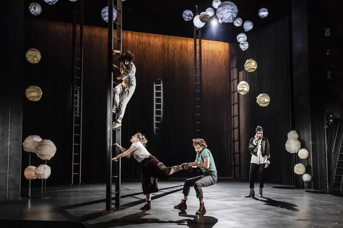 Boadicea Ricketts as Helena climbs one of five ladders on stage. Dawn Sievewright as Hermia tries to climb up towards Boadicea, while Ryan Hutton as Lysander grabs Dawn's leg. Bally Gill as Oberon stands to the right. White lanterns appear on the side and top of the stage.