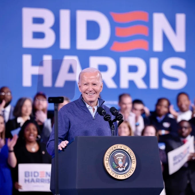 President Joe Biden delivers remarks behind a podium. 

There are supporters standing behind him with small Biden-Harris signs, and a large Biden-Harris sign hangs on a blue background behind him.