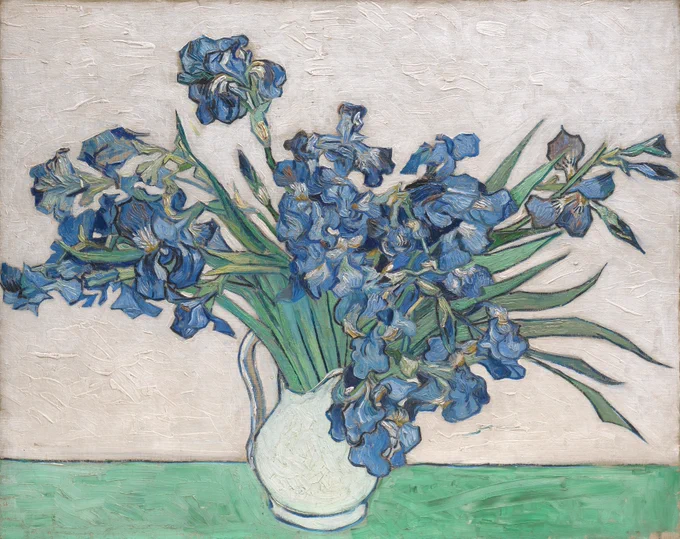 Blue irises vibrantly stretch out of a white vase sitting on a green table.