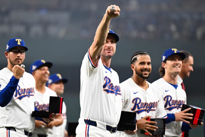 Max Scherzer raises his right fist to show his World Series ring to the crowd. He wears a white Rangers jersey with blue and red letters. 