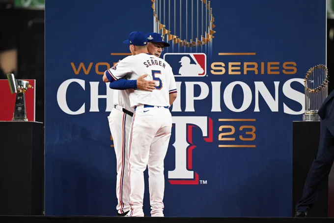 Corey Seager hugs Bruce Bochy on stage surrounded by the World Series and AL Championship trophies. They wear white Rangers jerseys with blue and red letters and numbers. 