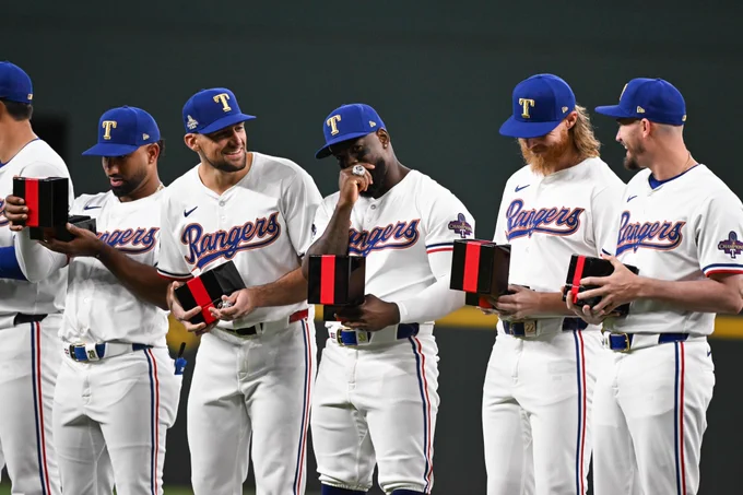 Adolis García laughs as he covers his mouth with his hand that has his World Series ring on it. Other Rangers players stand along the baseline during the ring ceremony. They wear white Rangers jerseys with blue and red letters. 