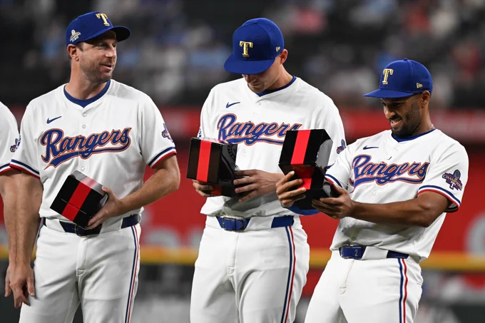 Corey Seager and Marcus Semien smile as they look inside a box to see their World Series rings. They wear white Rangers jerseys with blue and red letters. 