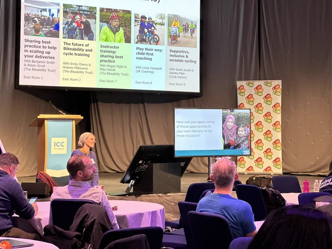 Delegates are sitting in a hall with a presentation screen displaying a woman wearing a hijab and the text: “How can you apply some of these approaches in your own delivery to be more inclusive?”