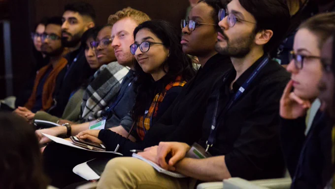 A group of people are sitting in a conference room looking at and listening to a speaker who is out of shot. 