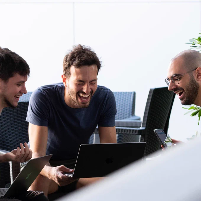 3 men looking at a laptop. The middle one is laughing.