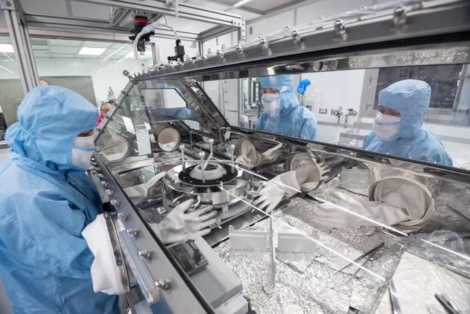 Three people dressed in blue clean lab suits interact with a large sample container within a hermetically sealed and windowed workspace.