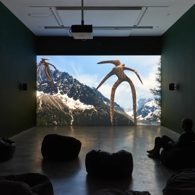 A viewer sits on a bean bag in an darkened gallery watching a video featuring an alien-like form in a landscape.