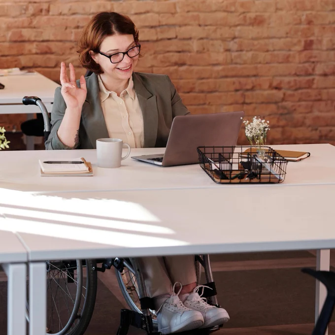 A woman sits at her desk in a wheelchair, smiling and waving at her laptop