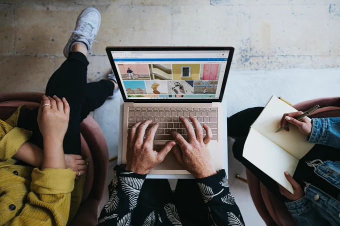 Photo by Windows on Unsplash depicts three unseen persons with the central person operating a laptop and the person on the right with a notepad and pen.