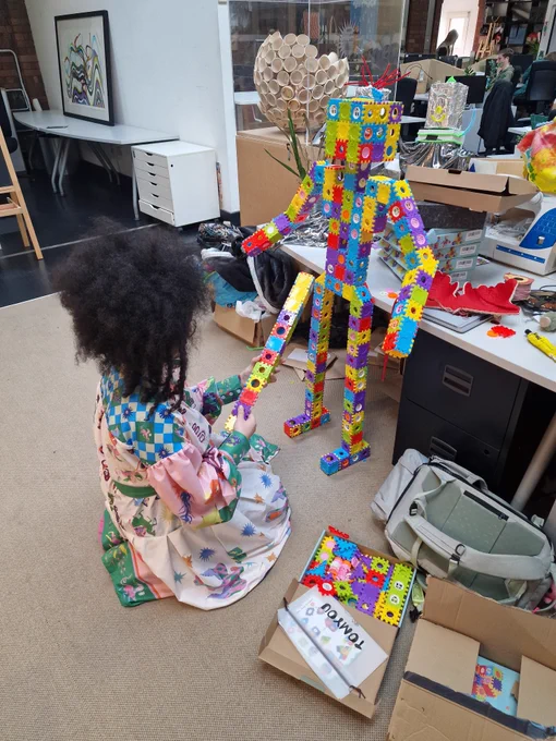 Image of a person in a colourful dress crouched on the floor of Pervasive Media Studio, and holding a long stack of colourful blocks. Right in front of them stands a sizeable humanoid figure constructed out of those same colourful blocks.