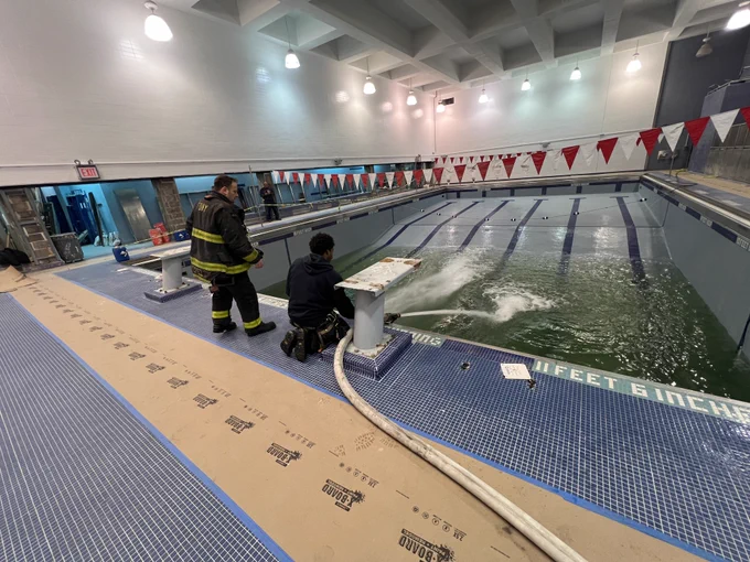 A firefighter is kneeling beside an empty pool spraying water through a fire hose. 