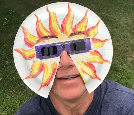A man smiles at the camera while weating eclipse glasses surrounded by a paper plate that has a crayon-colored sun covering most of his face like a mask, leaving his mouth and nose visible.