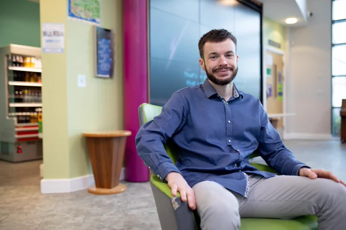 Smiling man Ross wearing a blue shirt and grey trousers with brown hair and a beard, sitting in a green chair in a colourful hospital waiting room with a bright pink pillar behind him in front of lime green walls.