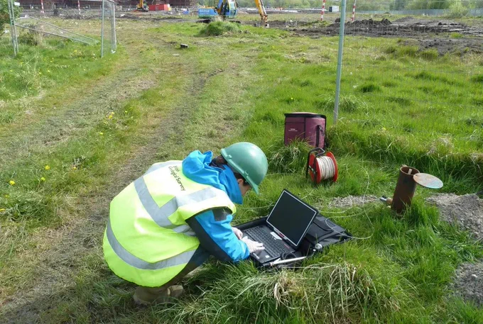A person doing ground investigations, crouching on the floor, looking into a laptop. A borehole sits in front of them. They are wearing full PPE. 