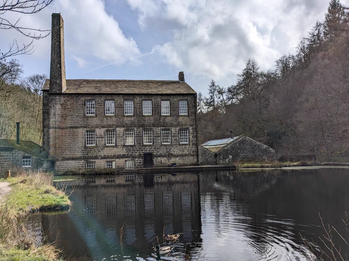 An old mill building with high chimney next to a pond with ducks, all in a valley surrounded by trees