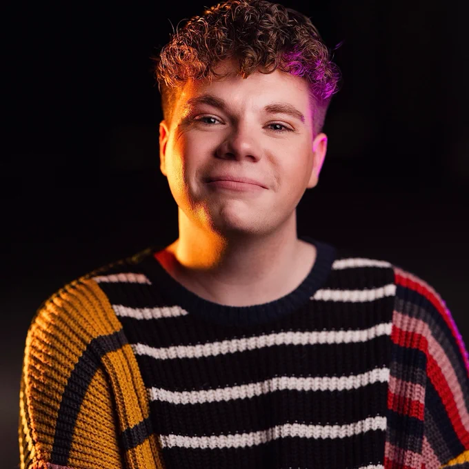 A man smiles whilst wearing a patterned jumper. He is lit by orange and purple light.