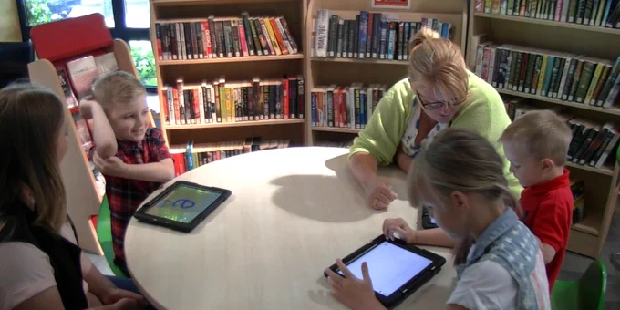 Image of two adults sitting at a round table with three children using iPads, in a library/bookshelves behind them