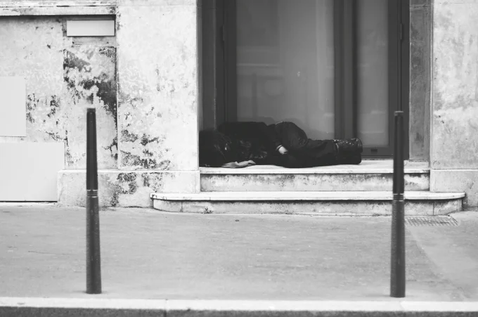 A black and white street photograph of a homeless person dress in dark clothing curled up asleep on the steps of a doorway. Stock image by Ev on Unsplash.