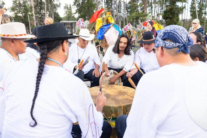 A group of Tribal singers and drummers sit in a circle outside with six tepees behind them. 