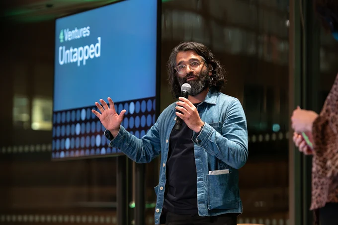 This image shows Vinay Solanki talking into a microphone on stage. He is wearing a denim jacket and black t-shirt. In the background you can see a screen which says 4 Ventures Untapped in white text.